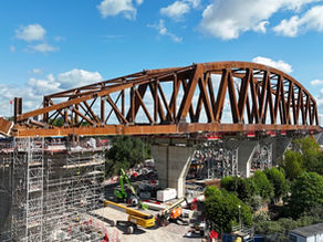 Rust-colored steel bridge arch under construction above scaffolding and cranes, with a bright blue sky and clouds overhead.