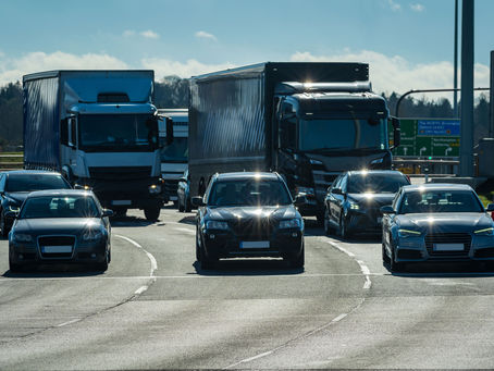 Cars and trucks drive on a sunlit highway, reflecting bright light. Road signs are visible in the background, with a clear blue sky above.