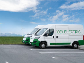 Two white-and-green electric vans parked by wind turbines, with 100% ELECTRIC on the side under a blue sky.