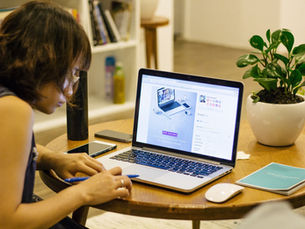 Woman writing on a wooden table with plant, notebook, and an open laptop nearby.