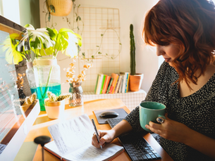 A woman with colored short hair, wearing a dotted top, copies data from her computer screen to her notebook by hand. She works in a home office brightly lit through a side window, with plants and a stack of books surrounding her workspace.