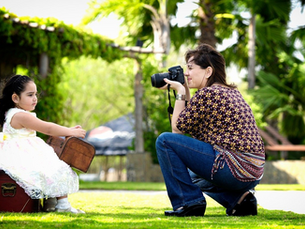 Woman photographs a little girl holding a suitcase on a sunny day. The girl is seated on another suitcase surrounded by greenery set against a house.