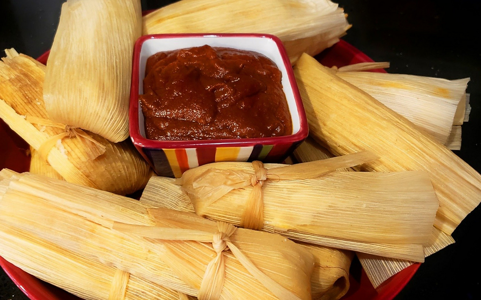A plate of tamales wrapped in corn husks surrounds a rainbow-colored bowl of red salsa.