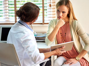 A lady doctor presents a tablet while in discussion with a thoughtful-looking lady patient in a sunlit office. 