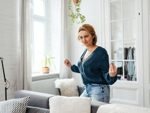 Woman in blue shirt and jeans dances happily in a bright living room with large windows, plants, and cozy decor.