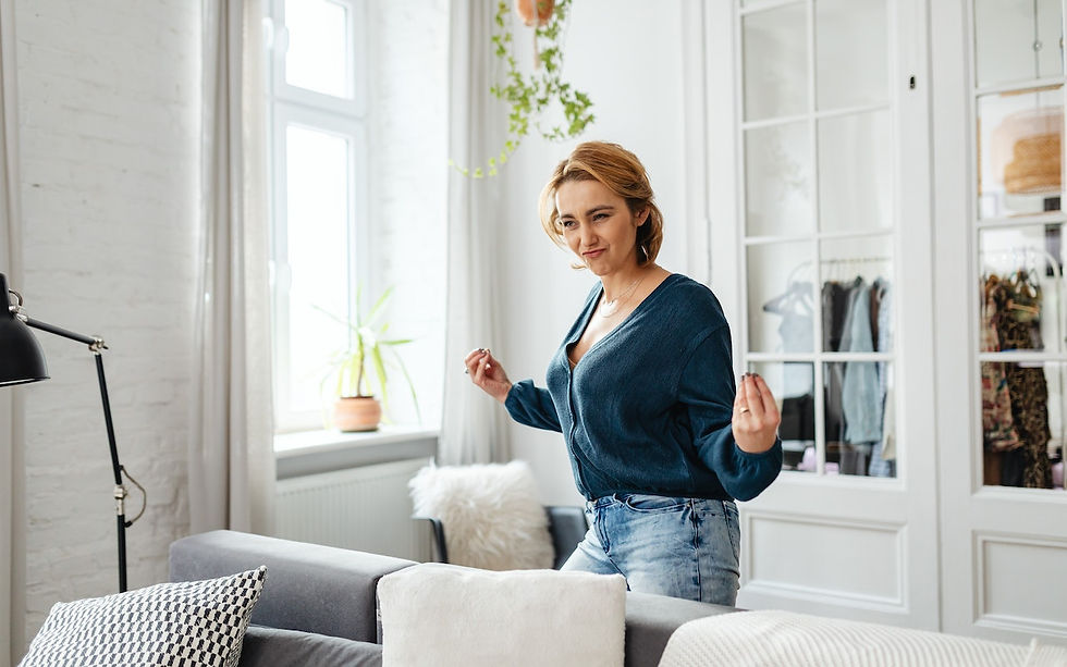 Woman in blue shirt and jeans dances happily in a bright living room with large windows, plants, and cozy decor.