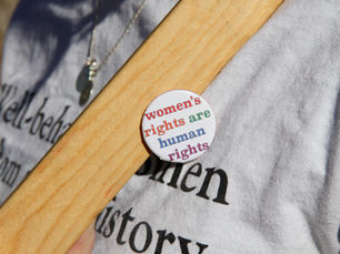 A badge reading "women's rights are human rights" is pinned to a person's shirt which has the (partially-hidden) printed message "Well-behaved women seldom make history."