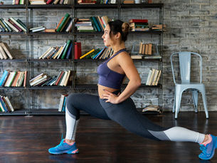A woman in stretchy exercise clothing does a lunge stretch against a background of brick walls and bookshelves.