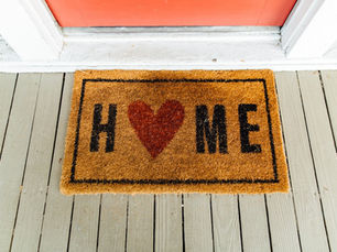 A mahogany-colored welcome doormat with "H❤️ME" text in black sits in front of a red door on a wooden porch.