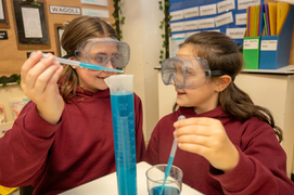 Two girls in goggles using pipettes and blue liquid for science experiment.
