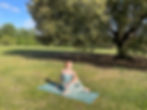 Emma in a seated spinal twist on her blue mat smiling in Greenwich Park. She is in front of a large tree.