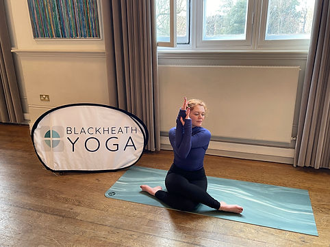 Emma sat in cowface pose with eagle arms in front of a blackheath yoga sign