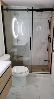 Modern minimalist bathroom featuring a black-framed sliding glass shower, a sleek floating wood vanity paired with marble-inspired walls and floors, a circular LED mirror, and matte-black fixtures including a white toilet.