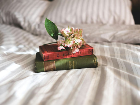 A stack of pink and white flowers sits on two books (The Isle of Wight Red Guide by Ward Lock and The New Forest by Rose C. de Crespigny and Horace G. Hutchinson) which rest on a bed covered in a white and gray striped comforter.