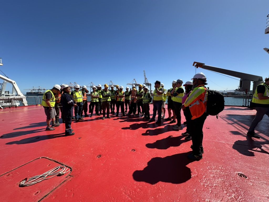 Crew in hard hats and hi-vis gathered for a briefing on a red deck