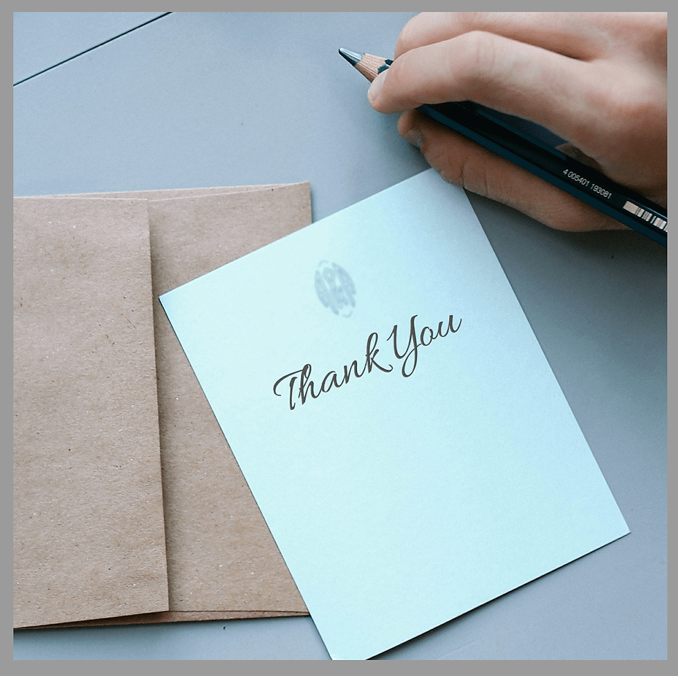 Hand holding a pencil near a light blue "Thank You" card with brown envelopes on a gray table. The mood is appreciative and thoughtful.