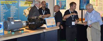 People in a booth at a trade show talk and gesture around display equipment. Blue informational panels and posters are visible in the background.