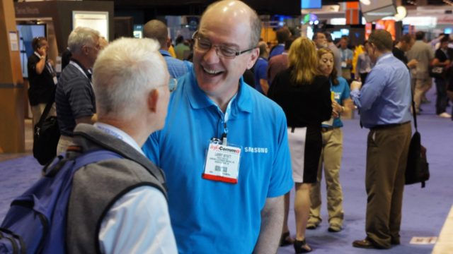 Two men talking at a busy tech convention. One wears a blue shirt with a name tag; both are smiling. Booths and attendees in background.