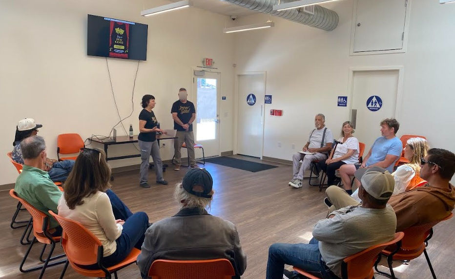 a group of people seated in a semi-circle, listening to a facilitator who is standing next to an assistant