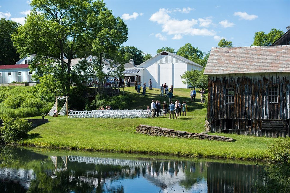 Outdoor wedding ceremony at a Vermont farm venue with white folding chairs set on a hillside lawn, a reflective pond, and a large white event barn, Scott Farm Orchard style wedding grounds