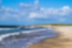 Strand am kyst til Kyst Stien mit sanften Wellen, blauer Himmel und Wolken. Grüne Dünen im Hintergrund. Entspannende, friedliche Küstenlandschaft.