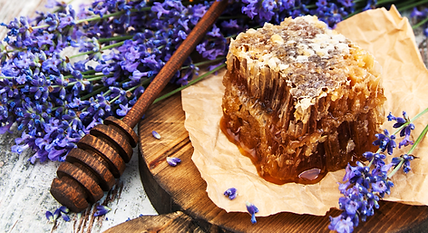 A piece of honeycomb and bundles of lavender arranged on a wooden cutting board next to a honey dipper.