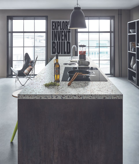 Side view of kitchen with cooking on island in Schuller German kitchen with dark textured wood, terrazzo worktops, and concrete floors