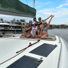 Five women relax on the upper deck of a catamaran in Fort Lauderdale, Florida, during a bachelorette party. The bride, wearing a swimsuit printed with the groom’s face, sits in the center as friends smile and pose around her