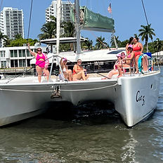 A group of women in swimsuits lounge on the front deck of a catamaran in Fort Lauderdale, Florida, during a bachelorette party. Some sit on the trampoline net while others pose near the bow, with waterfront homes and palm trees in the background