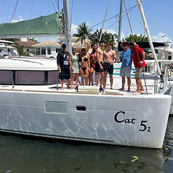 A mixed group of men and women stand on the deck of a catamaran named 'Cat 5-1' in Fort Lauderdale, Florida, during a bachelorette party. They pose together in swimsuits under a sunny sky, with palm trees and waterfront homes in the background
