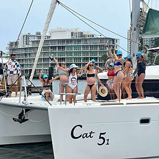 A group of women in swimsuits and matching light blue caps dance and pose on the deck of a catamaran named 'Cat 5-1' in Fort Lauderdale, Florida, during a bachelorette party. The bride wears a swimsuit printed with the groom’s face, and the city skyline is visible in the background
