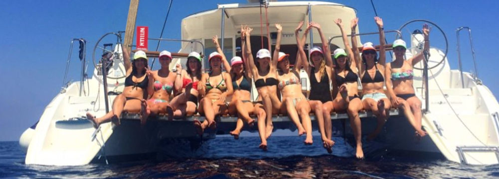 group of women in swimsuits sit side by side on the stern of a catamaran, legs dangling over the water. They wear colorful caps and raise their arms in celebration, smiling under a clear blue sky