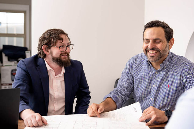 Two men smiling while looking at a document together at a table.