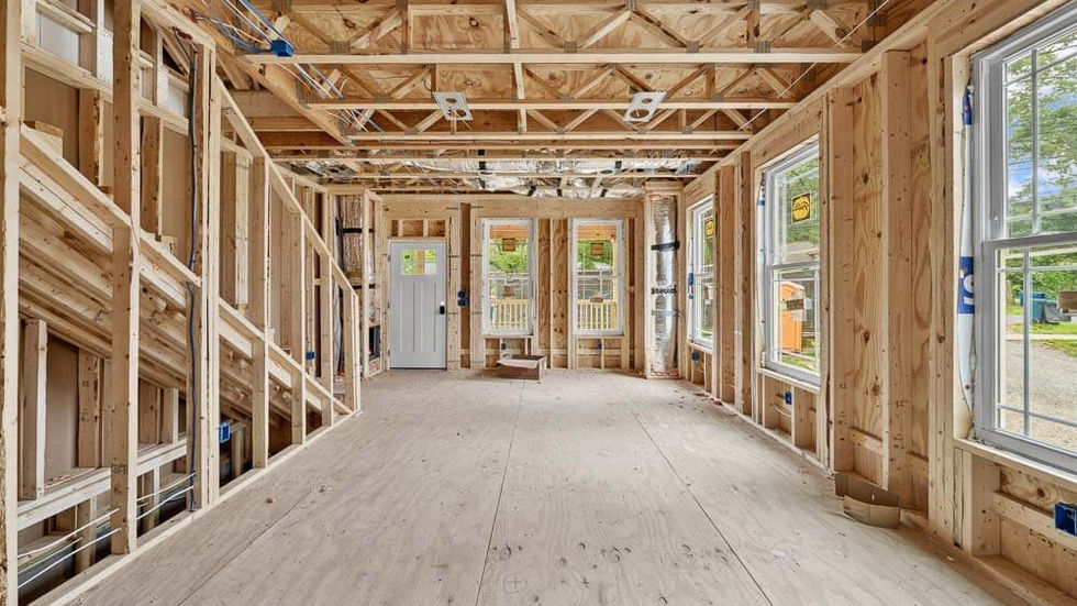Interior view of a house under construction, showing exposed wood framing, unfinished flooring, installed windows, and rough electrical work before drywall installation.