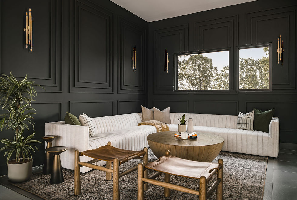 Modern lounge with dark paneled walls, cream sectional, round wood table, leather stools, plants, and windows showing trees.