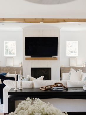 Modern living room with white couches, blue chairs, a large TV above a fireplace, and wooden accents. Bright, airy ambiance with stairs.
