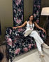 Smiling woman in white outfit sits on a black floral sofa beside a lamp, wearing a lanyard in a cozy room.