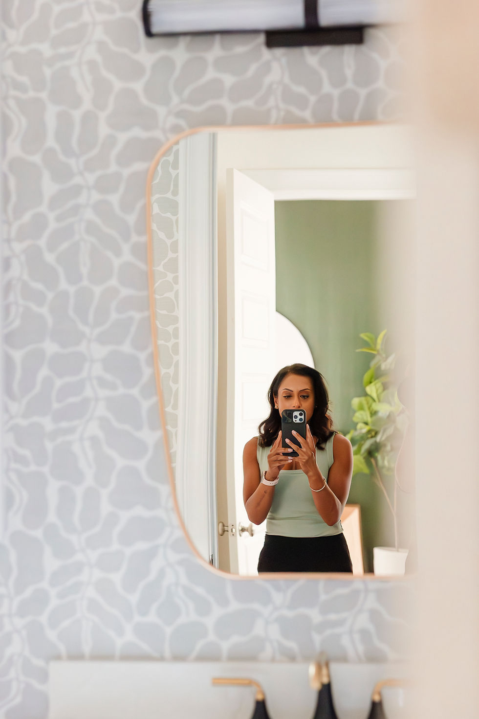 Woman taking a mirror selfie in a bathroom with a patterned wall. She's wearing a green top and black pants. Mood is neutral.