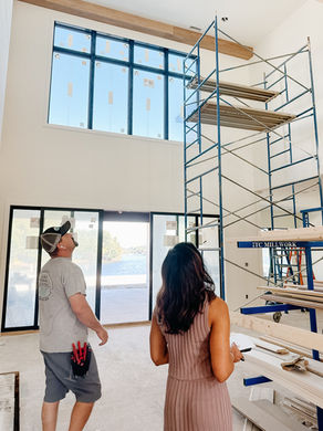 Two people observe a tall indoor scaffold near large windows. Bright setting with construction tools visible, overlooking a scenic lake view.