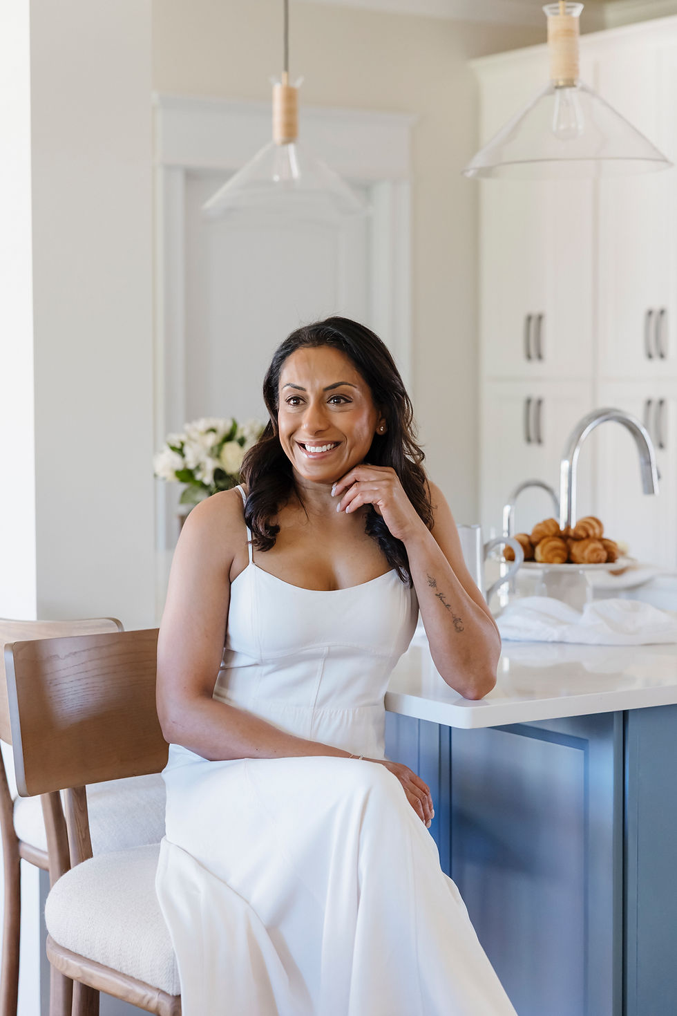 A woman in a white dress sits smiling at a kitchen counter with croissants and flowers. Bright, minimal décor. Mood is cheerful.