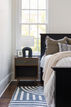 Bedroom with a modern nightstand, white lamp, candle, and gray pillow. Large window shows green leaves. Striped rug enhances coziness.