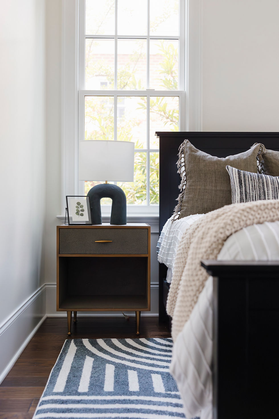 Bedroom with a modern nightstand, white lamp, candle, and gray pillow. Large window shows green leaves. Striped rug enhances coziness.