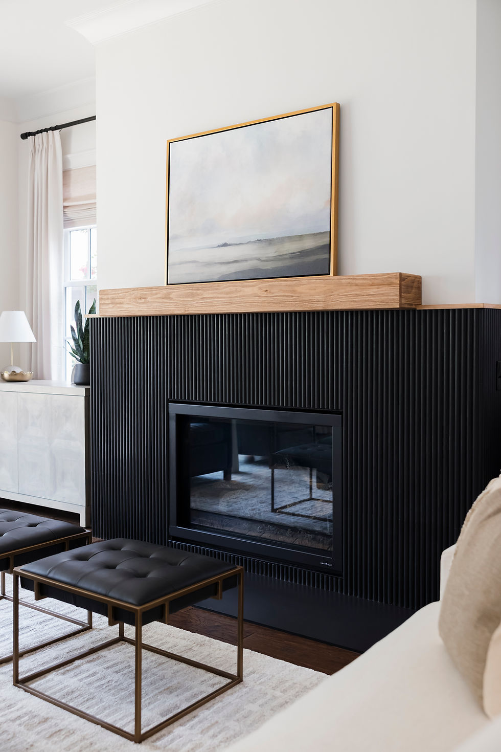 Modern living room with a black fireplace, a landscape painting above, two black stools, a white rug, and light curtains. Minimalist decor.