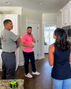 Three adults talk in a bright white kitchen, one in pink holding a phone, with a small tape measure on the counter.