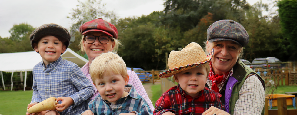 Two staff members and children, dressed as farmers, pose on a wooden gate outdoors with big smiles.