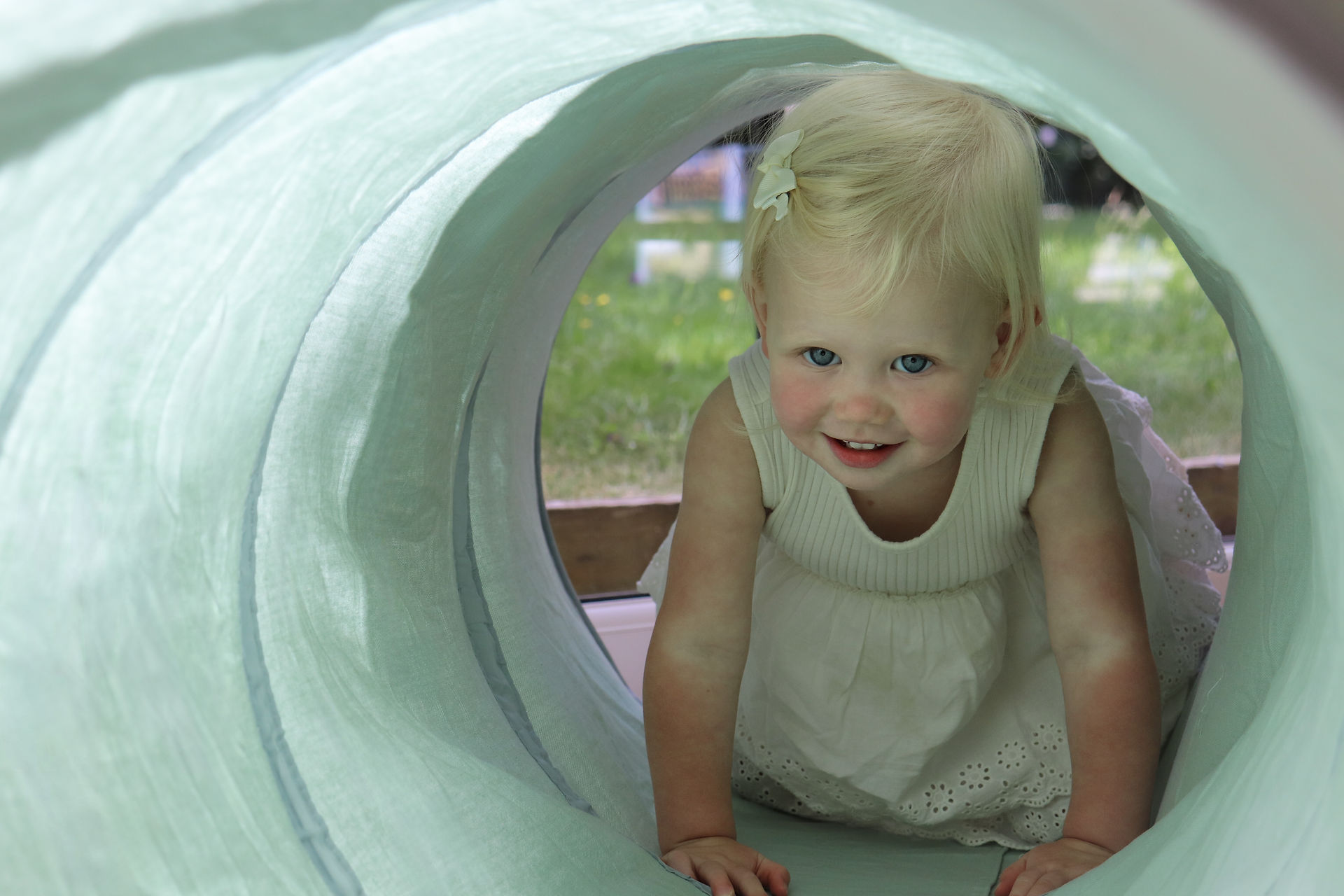 A smiling toddler explores a fabric play tunnel, building confidence and motor skills through movement and playful discovery.
