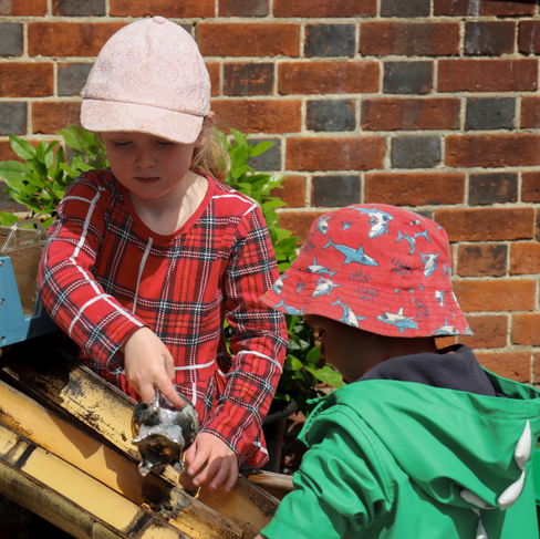 Two children in outdoor play at a Reading nursery. The girl pours water while the boy watches, exploring water play.