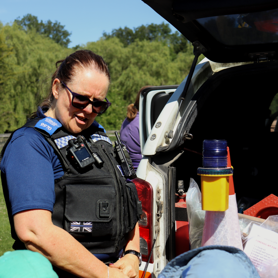 Female police officer explaining equipment to children by an open police vehicle during a sunny nursery event.