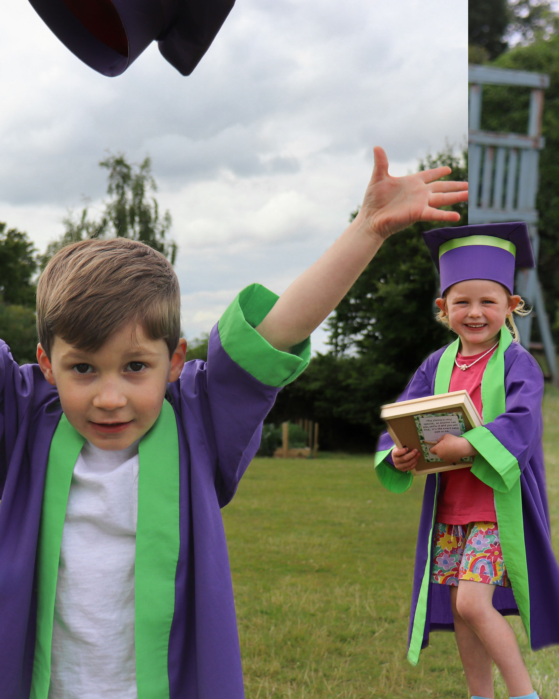 Two children in purple and green graduation gowns celebrate outdoors. One throws his cap while the other smiles.