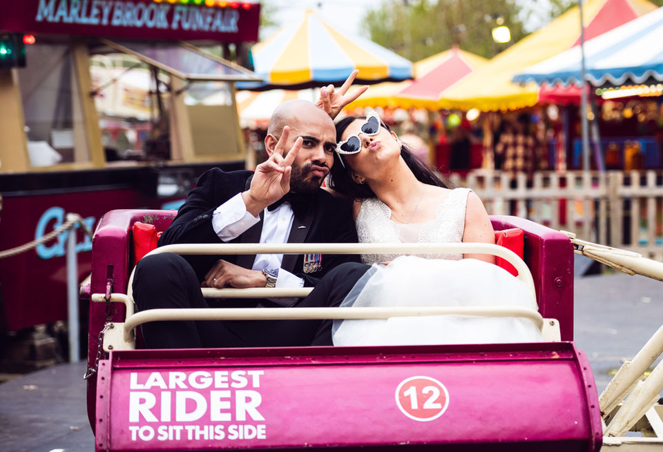 Photograph of a newly married couple on the fun fair rides at their wedding venue in Kent Bride wearing her large white wedding dress and the groom smartly dressed in black suit and sun glasses photo taken by wedding photographer Dover Media Group